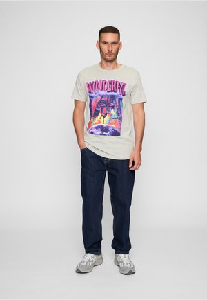 Mand stående med hænderne i lommerne iført en beige T-shirt med teksten "Wonderful", mørke jeans og grå sneakers foran en ensfarvet baggrund.