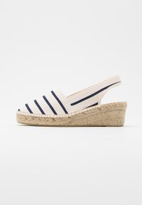 Béžová espadrilková sandála na klínu s námořnickými modrými vodorovnými pruhy a popruhem na patě na bílém pozadí.