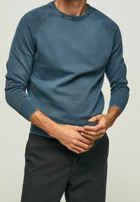 Muž v modrém pleteném svetru s dlouhým rukávem a černých kalhotách, s rukama sepnutýma v pase na jednobodovém pozadí.