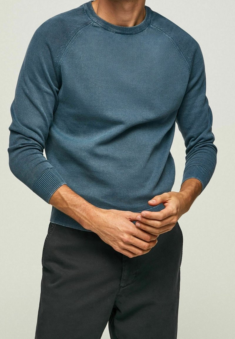 Muž v modrém pleteném svetru s dlouhým rukávem a černých kalhotách, s rukama sepnutýma v pase na jednobodovém pozadí.