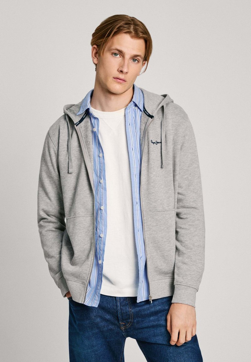Felpa grigia con zip e lacci, indossata sopra una t-shirt bianca e una camicia a righe blu chiaro con bottoni, abbinata a jeans blu scuro.
