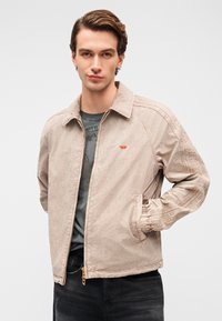 Φωτεινό εκρού μπουφάν με λεία υφή, διαθέτει φερμουάρ, ριμπ μανσέτες και μικρό κόκκινο λογότυπο στο στήθος.