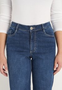 Närbild av en person som bär högmidjade blå denimjeans med knapp- och dragkedjeslutning, i kombination med en vit långärmad topp.