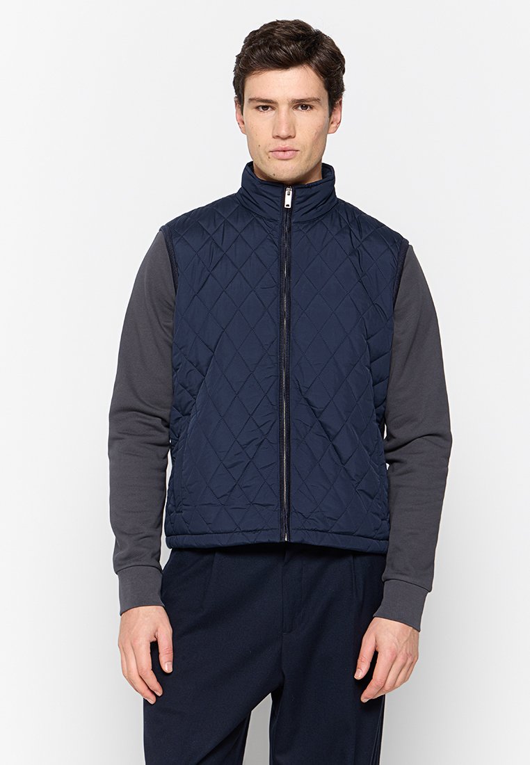 Gilet matelassé marine avec un col haut et une fermeture éclair à l'avant, présentant un motif texturé. Porté par-dessus une chemise à manches longues grise et un pantalon foncé.