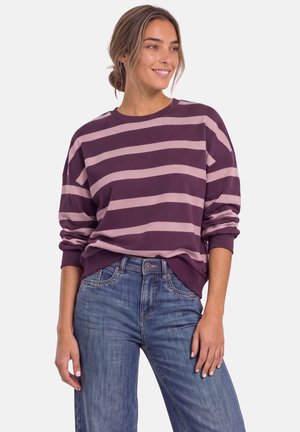 Kvinde smiler iført en lilla og lyserød stribet sweater med opkørte ærmer og blå jeans mod en ensfarvet baggrund.
