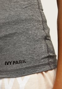 Gråt fitted linne i texturerad tyg, med svart "IVY PARK"-logotyp vid fållen, med synliga sömdetaljer.