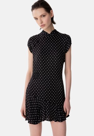Donna che indossa un vestito nero a maniche corte con pois bianchi, un orlo plissettato e una scollatura alta su uno sfondo neutro.