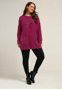 Pull oversized magenta avec des motifs de nœud en sequins, manches longues, encolure ronde, associé à un pantalon noir ajusté et des bottines.