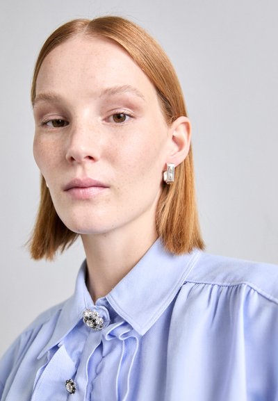 Blue collared shirt with pleated details and decorative buttons. Silver geometric earrings accent the look. Smooth texture, minimalist design.