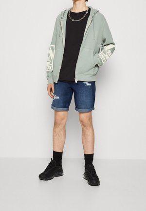 Haut zippé vert avec des manches imprimées, t-shirt noir, shorts en denim déchirés et baskets noires. Les accessoires comprennent un collier en chaîne argenté.