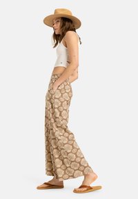 Pantaloni largi cu un model maro și un design floral, confecționați dintr-un material ușor. Purtați cu un top alb scurt și sandale maronii.