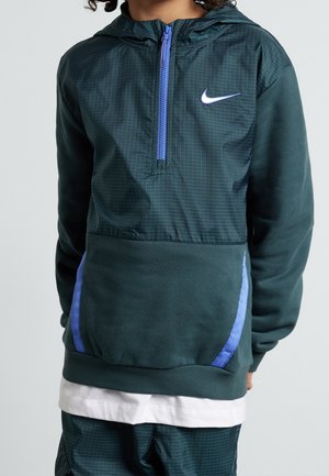 Enfant portant un sweat à capuche Nike vert foncé avec une demi-fermeture éclair bleue et des accents de poche, associé à un pantalon à motif quadrillé assorti et un sous-vêtement blanc.