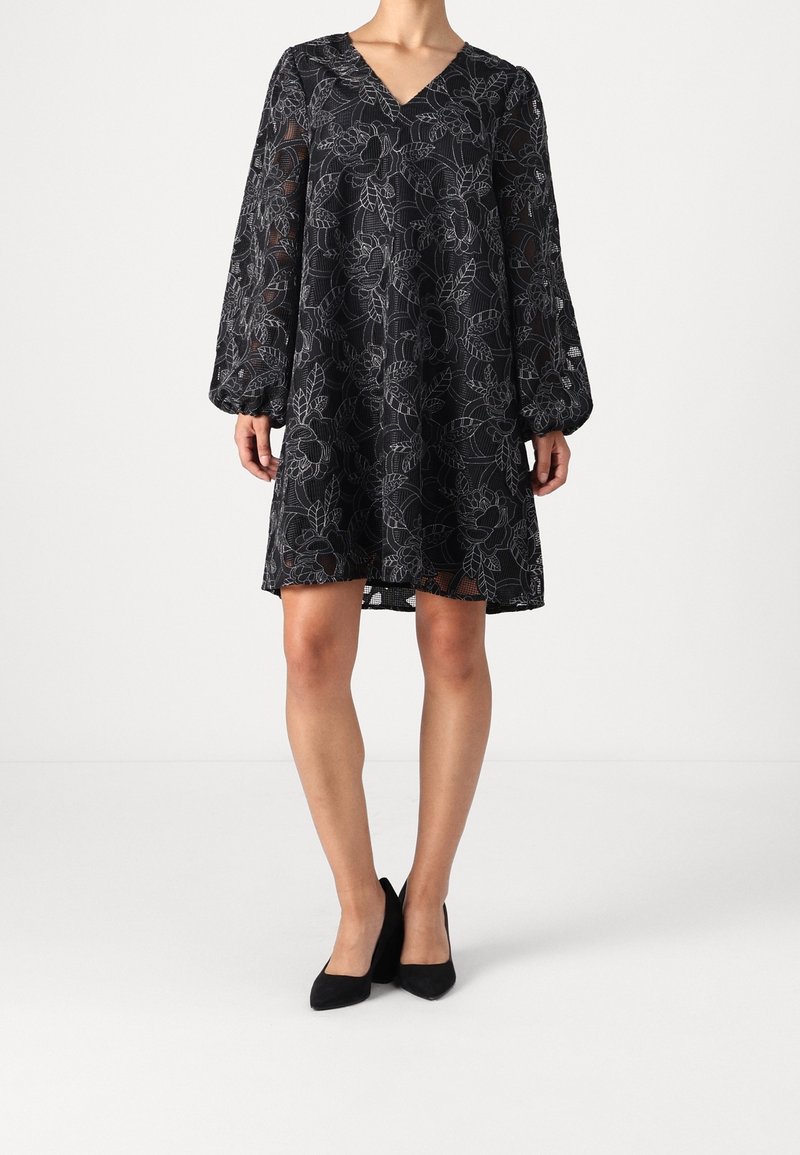 Femme portant une robe noire mi-longue avec des manches longues transparentes et des motifs floraux blancs, associée à des escarpins noirs pointus.