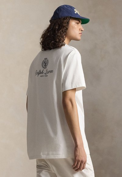 Junge Person mit lockigem Haar trägt ein weißes Ralph Lauren T-Shirt und eine blaue Kappe, seitlich stehend vor einfarbigem beigem Hintergrund.