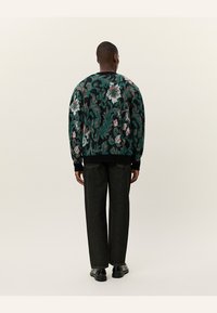 Pull en maille verte texturée avec des motifs floraux rose et blanc, poignets côtelés noirs, associé à un pantalon droit noir.