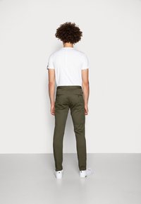 Chino verde oliva con vestibilità slim, caratterizzati da una texture liscia, tasche posteriori con cerniere, abbinati a una maglietta bianca a maniche corte.