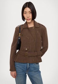 Brown checkered jacket with structured shoulders, front zipper, belt detail, and two side pockets. Worn over blue jeans.