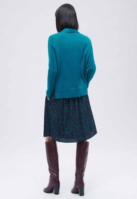Teal gebreide trui gecombineerd met een donkere bloemenjurk. Het model draagt bruine kniehooglaarzen. De outfit heeft een platte kraag en geribde manchetten.