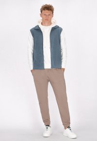 Blaues Fleece-Weste über einem weißen Hoodie, kombiniert mit beigen Jogginghosen und weißen Sneakers. Auffällige Merkmale sind der Reißverschluss vorne und die Taschen.