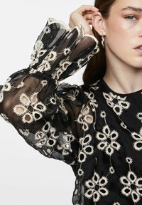 Sort gennemsigtig bluse med broderede hvide blomster mønstre. Har puffede ærmer med elastiske manchetter og runde udskæringer i stoffet.