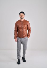 Veste en cuir marron avec un col montant, une fermeture éclair et une poche latérale. Le mannequin porte un pantalon gris et des bottines noires.