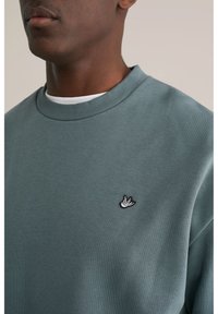 Zelena majica z raglanskim ovratnikom z rebrastim vzorcem in vezenim logotipom ptice na prsih. Tkanina je videti mehka in teksturirana.