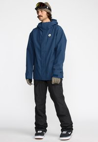 Veste imperméable navy avec capuche réglable, fermeture éclair à l'avant et logo. Associée à un pantalon cargo noir et des gants marron. Des lunettes de neige sur la tête.