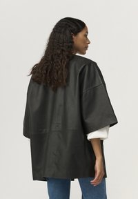 Fekete bőr, oversized dzseki sz�éles ujjakkal, sima textúrával és letisztult dizájnnal. Fehér ing és kék farmer felett viselve.
