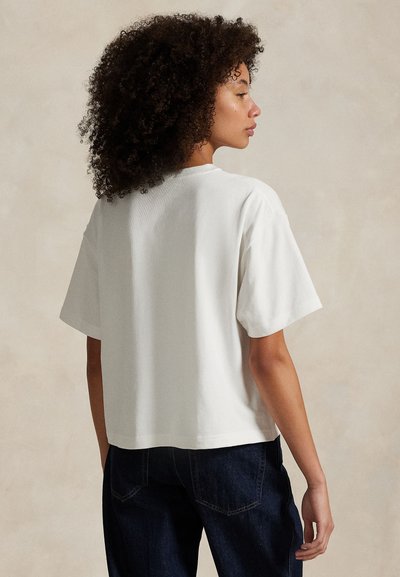 Kobieta z kręconymi włosami widziana od tyłu, ubrana w luźną białą koszulkę z krótkim rękawem i ciemne jeansy na neutralnym tle.