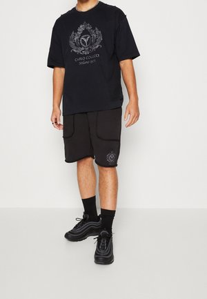 Homme portant un t-shirt Carlo Colucci noir et un short assorti avec des logos imprimés, des chaussettes noires montantes et des baskets noires, debout sur un fond uni.