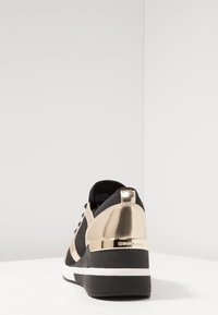 Buty sportowe z czarną siateczkową cholewką, złotymi metalicznymi akcentami oraz grubą czarną podeszwą. Widok z tyłu podkreśla kontrastujące faktury i kształty.