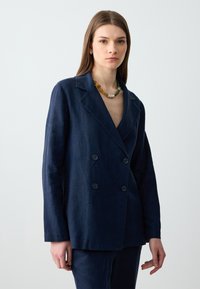 Granatowa lniana klasyczna marynarka dwurzędowa z wyłogami, z dwoma guzikami i luźnym krojem. Modelka nosi naszyjnik z wielokolorowych koralików.