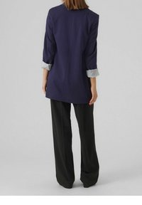 Donkerblauwe blazer met opgerolde mouwen met strepen, gecombineerd met wijduitlopende zwarte broek. Eenvoudige, op maat gemaakte silhouet, gladde stof.