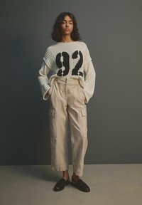 Gruba biała sweter z czarną grafiką "92", w połączeniu z beżowymi spodniami cargo z przednimi kieszeniami i podwiniętymi mankietami oraz czarnymi butami wsuwanymi.