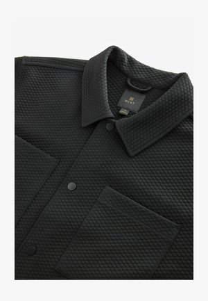 Zwarte gewatteerde jas met een omklapbare kraag, knopen aan de voorkant en een borstzak. De stof heeft een gestructureerd, diamantpatroon. Merkenlabel zichtbaar.