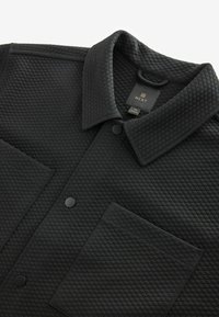 Gestepptes schwarzes Shirt-Jackett mit spitzem Kragen, Fronttasche und schwarzen Druckknöpfen. Texturierte Oberfläche und Etikett im Kragen sichtbar.