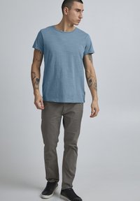 Lyseblå t-skjorte med kort erm og rund hals, kombinert med grå bukser og svarte joggesko, båret av en person med tatoveringer.
