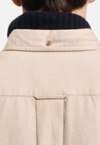 Camisa de espiga beige con cuello de canalé azul marino. Presenta un botón en la parte posterior y un detalle de pliegue para mayor textura.
