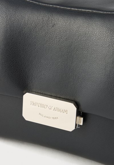 Crna kožna torba s metalnom srebrnom pločicom na kojoj je utisnut naziv marke "Emporio Armani" i "Milano 1981". Glatka tekstura.
