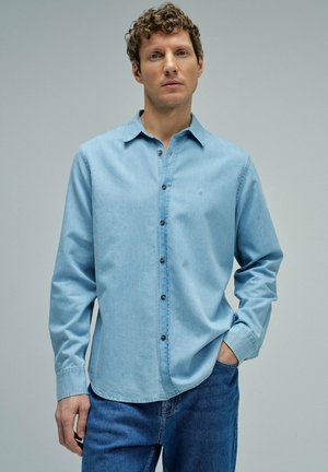Homme aux cheveux bouclés portant une chemise bleu clair à boutons et un jean bleu, debout devant un fond gris uni.