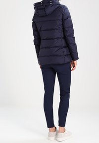 Donna che indossa una giacca trapuntata blu navy con cappuccio, pantaloni slim blu navy e sneakers beige chiaro, in piedi di spalle su uno sfondo bianco.