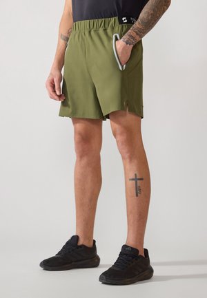 Oliven grønn idrettsshorts med elastisk midje, sidelommer og splittdetaljer ved kanten. Brukt med svarte joggesko.
