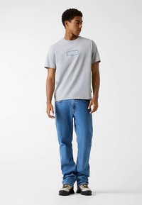 Grijs katoenen T-shirt met korte mouwen, voorzien van een blauw logoplaatje, gecombineerd met blauwe spijkerbroek en robuuste grijze en zwarte schoenen.