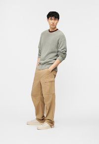 Νεαρός άνδρας που φοράει γκρι πλεκτό πουλόβερ και μπεζ παντελόνι φορτίου, στέκεται με τα χέρια στις τσέπες μπροστά σε απλό λευκό φόντο.