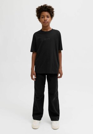 Sort bomulds T-shirt med rund hals og diskret logo, parret med sorte bukser og hvide sneakers, mod en neutral baggrund.