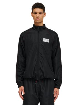 Young man wearing a black zip-up jacket with red piping and a white rectangular patch on the left chest, paired with matching black pants.