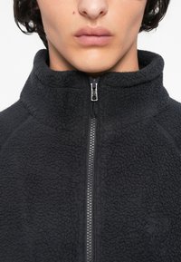 Primo piano di una persona che indossa una giacca in pile nera con texture e zip, con un colletto alto, parzialmente chiusa, che mostra le labbra e il mento.