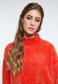 Pulover portocalie, texturat, cu un guler înalt și ribonat. Țesătura pare moale și pufoasă, având o croială lejeră și un model de cusătură vizibil.