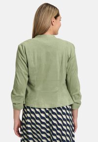 Veste en suède vert clair, à la texture douce, avec des manches trois-quarts et une silhouette ajustée, associée à une robe à motifs avec des lignes ondulées.