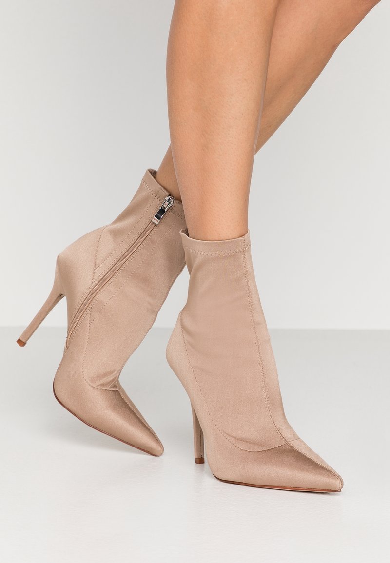 Piernas de mujer que llevan botas de tacón alto, de punta y tobillo, en satén beige con cremalleras laterales, sobre un suelo blanco.